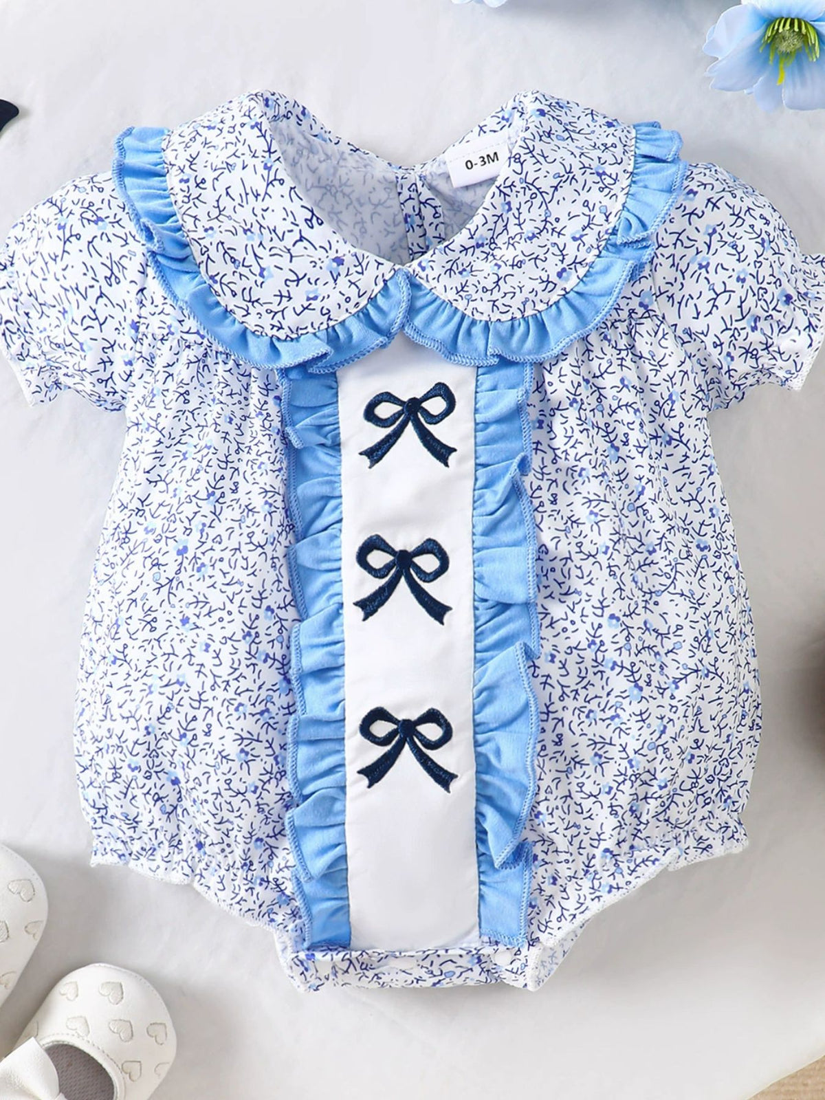 Baby one-piece with floral print, ruffle collar, and three embroidered bows on pastel blue trim.