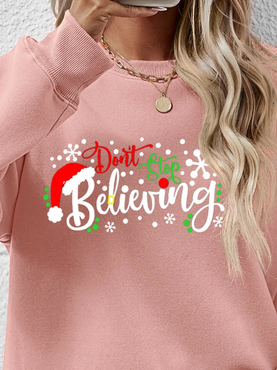 Blush pink long sleeve sweater with "Don't Stop Believing" holiday graphic and Santa hat detail.