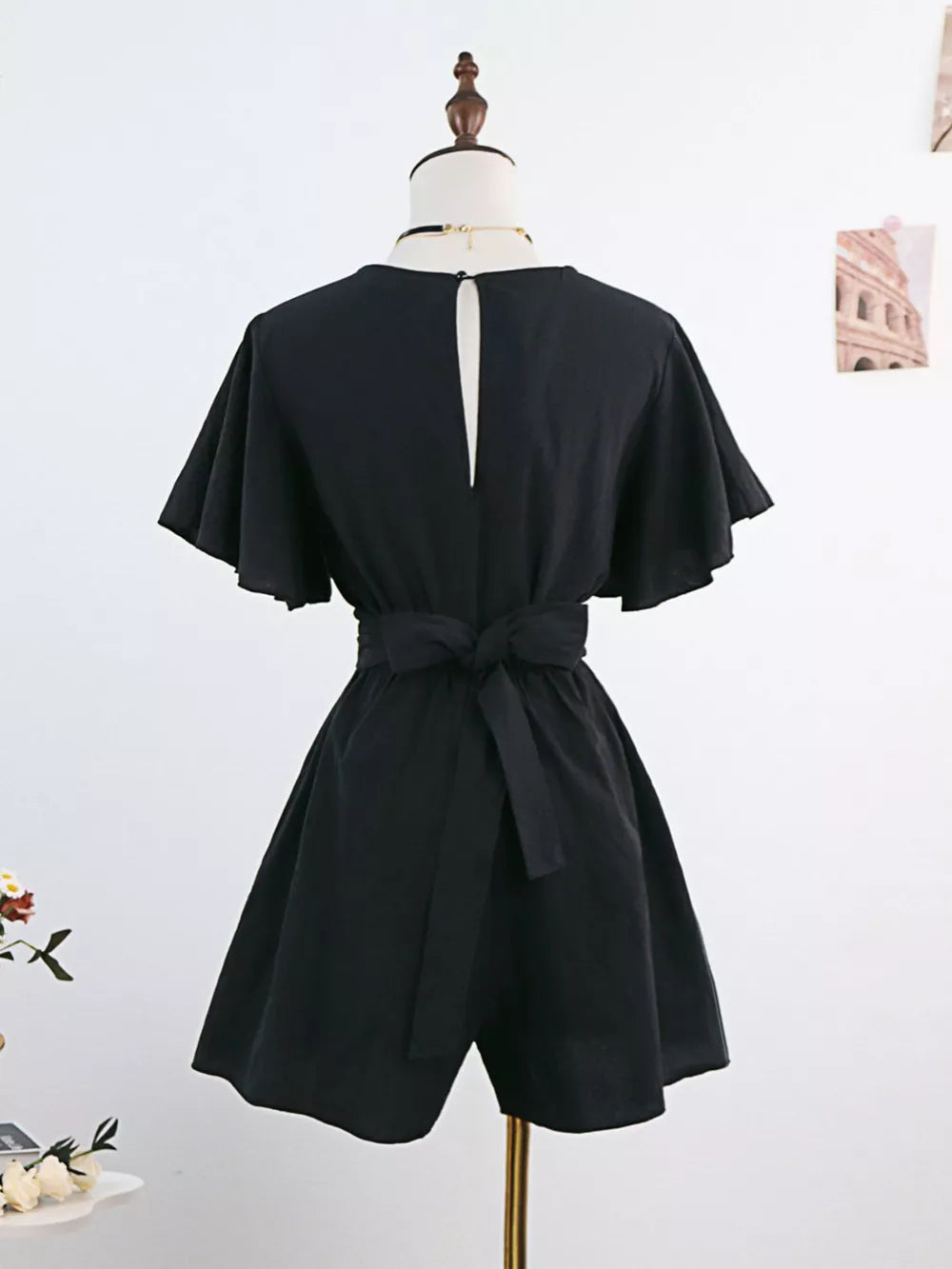 Black cotton romper with flutter sleeves, tie waist, and back keyhole detail.