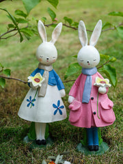 Floral resin rabbit figurines in blue and pink coats, holding flowers.