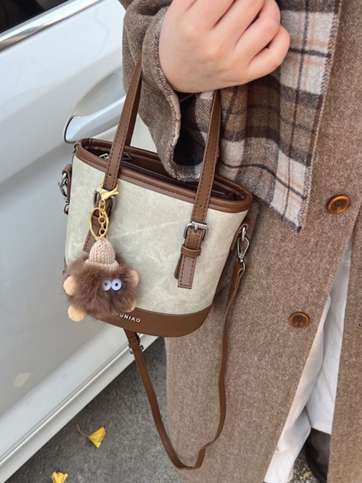Premium two-tone bucket crossbody bag in beige canvas with brown trim and fuzzy charm.
