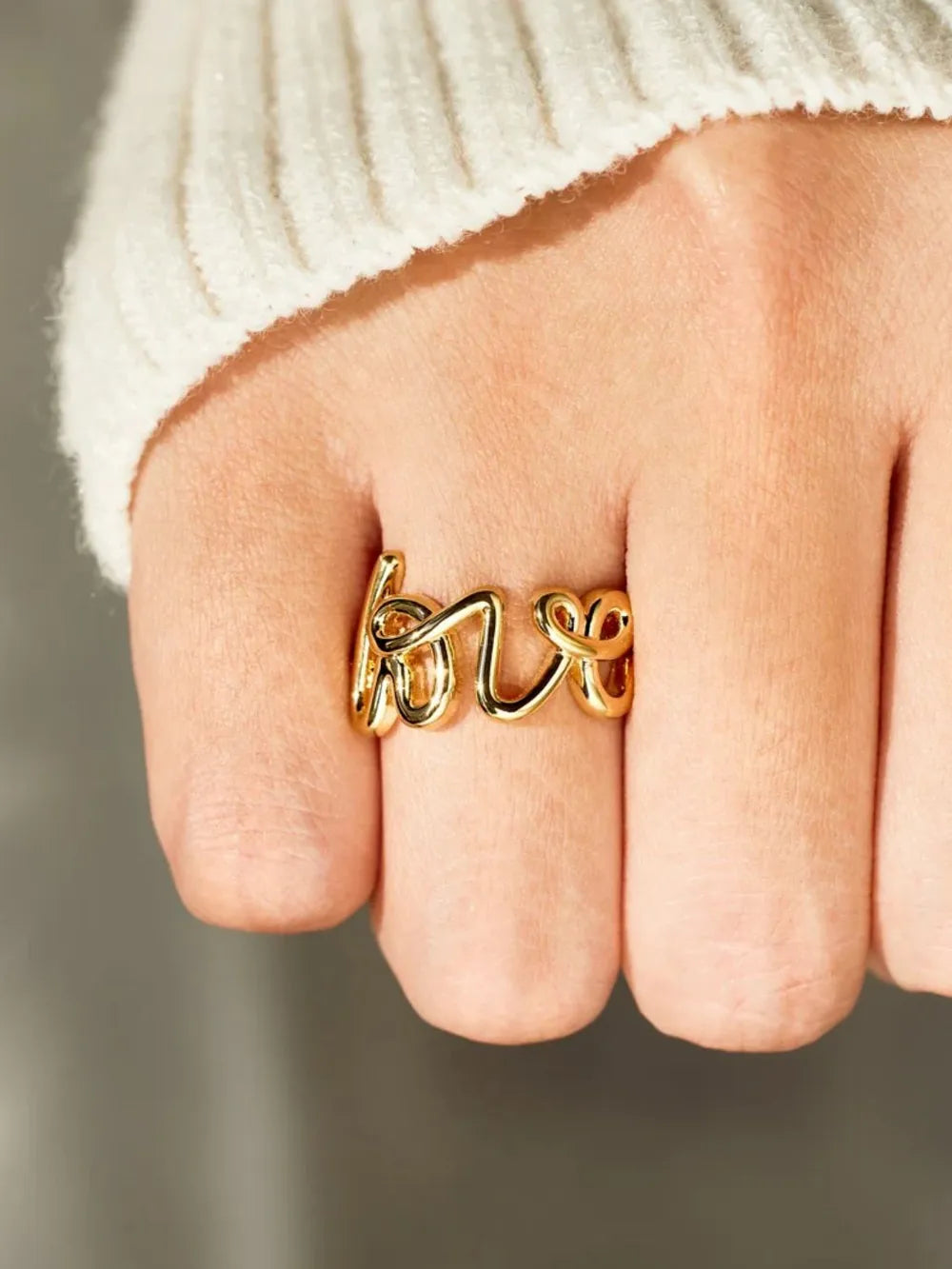 Gold scripted love ring on a finger, styled with a white sweater.