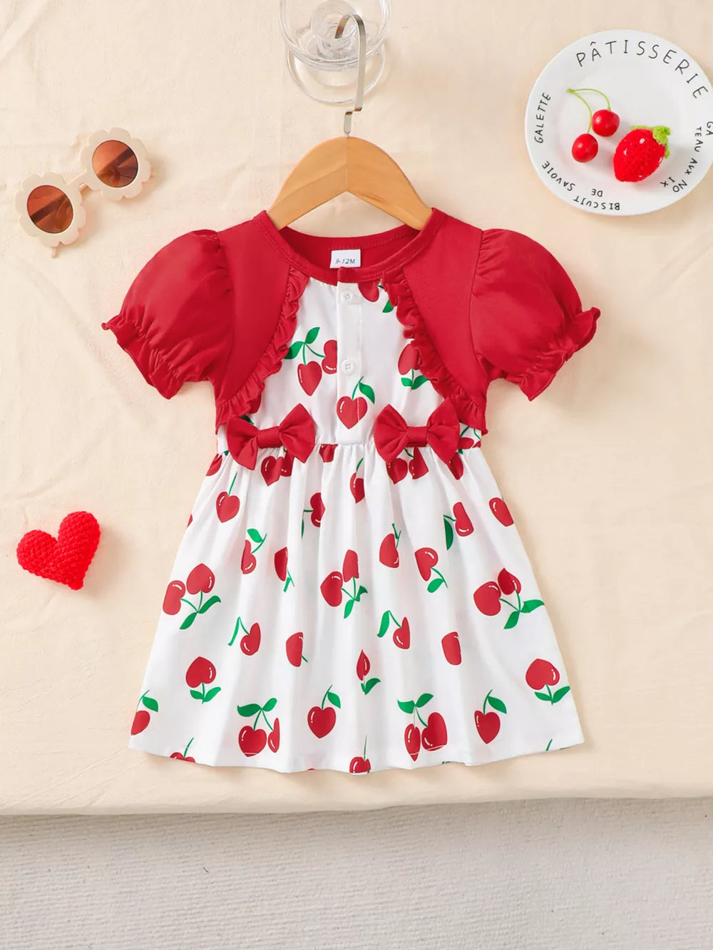 Baby dress with red cherry print, ruffled neckline, short sleeves, and bow details.