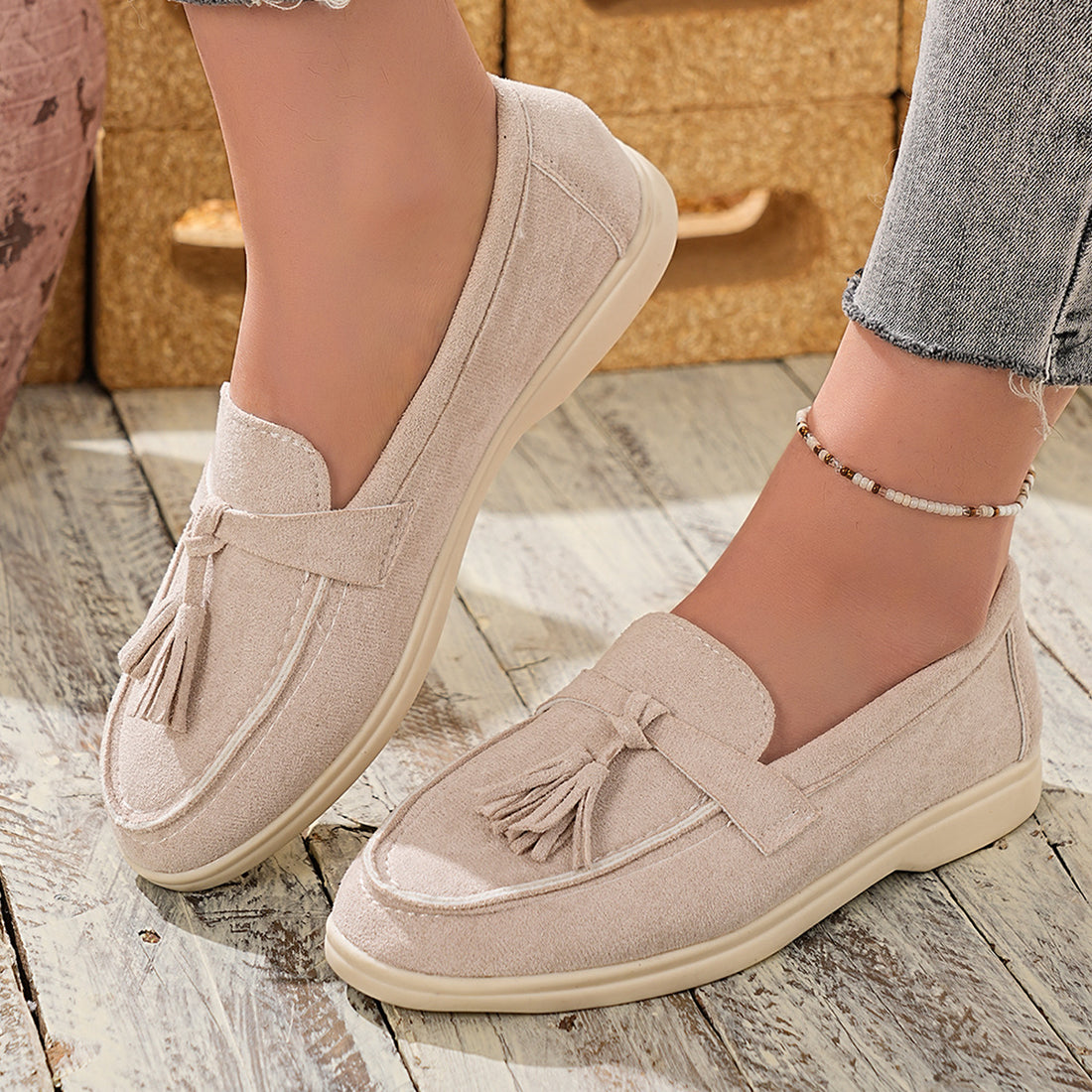 Beige faux-suede slip-on loafers with decorative tassels and contrast stitching.