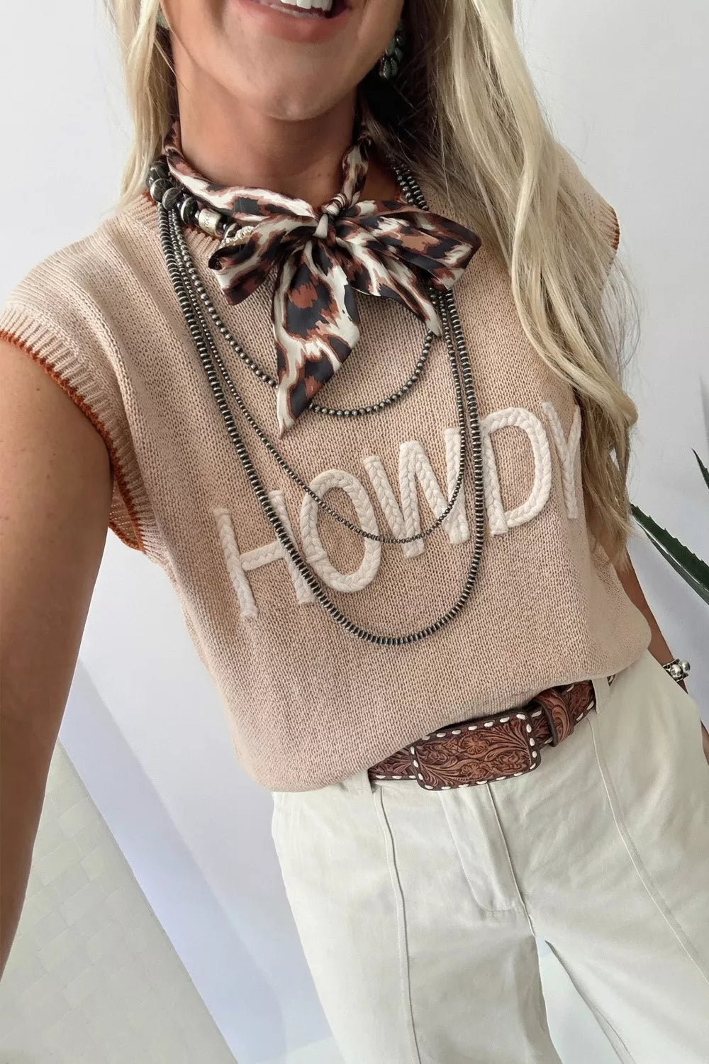 Howdy Textured Knit Sweater Vest in beige with white braided text, sleeveless, crew neck, and orange trim.