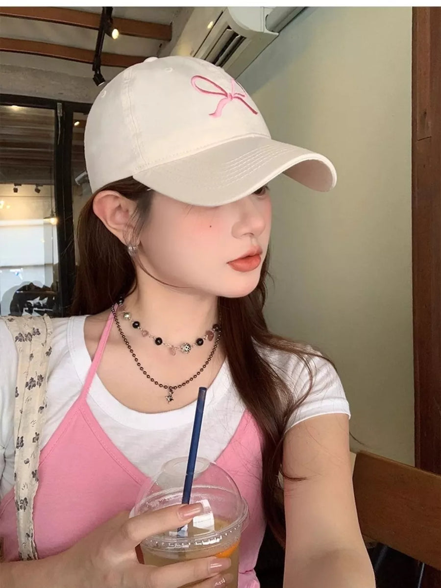 White baseball cap with pink bow embroidery, styled with a pink cami and layered necklaces.