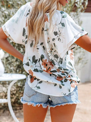 White floral print v-neck blouse with short sleeves and tie detail, worn with denim shorts.