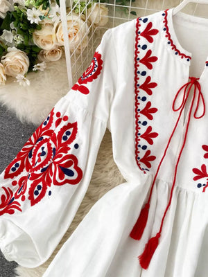 White maxi dress with red and blue embroidery, balloon sleeves, and tassel ties.