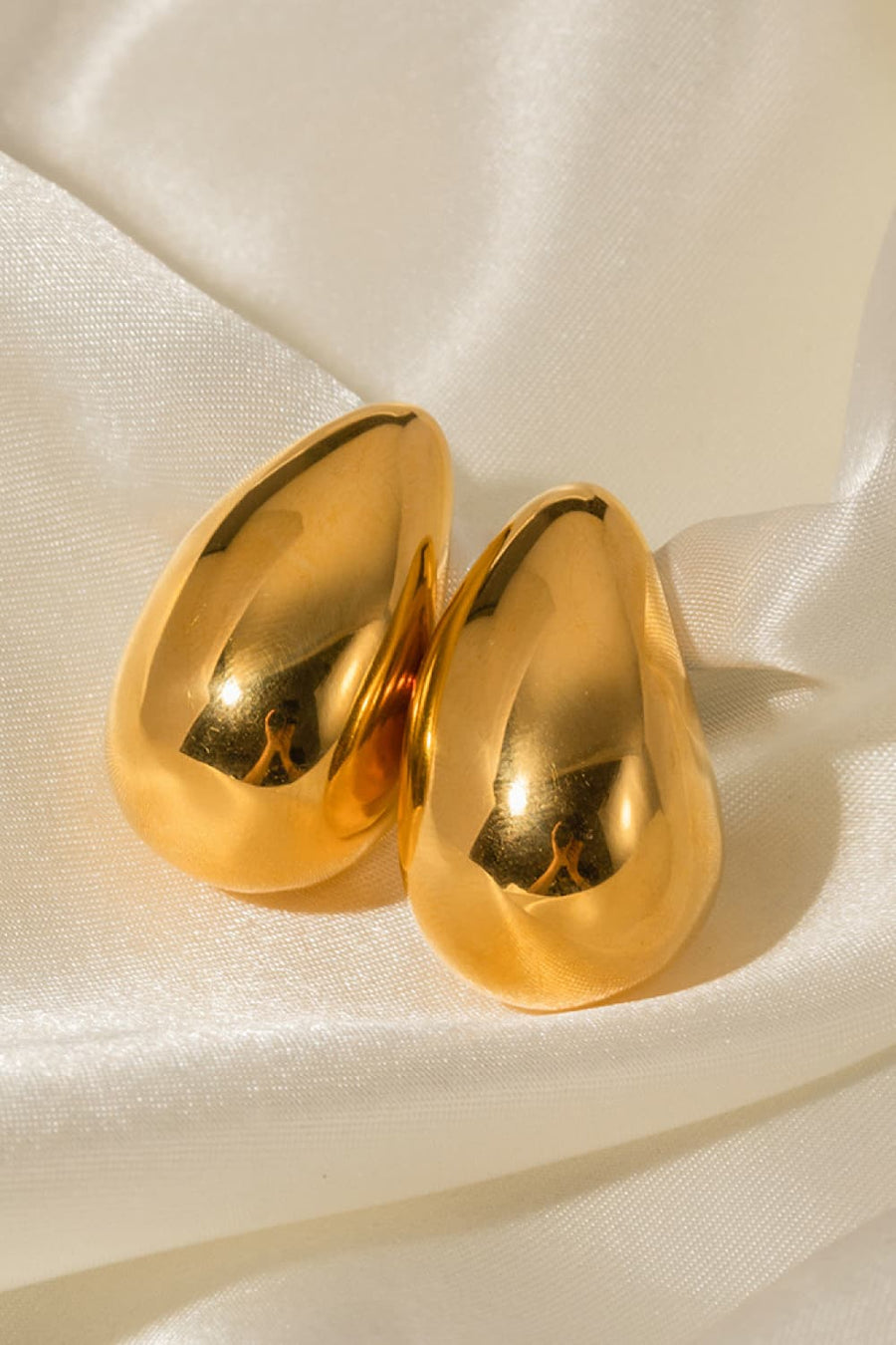Sculptural gold teardrop stud earrings with a polished finish on satin fabric.