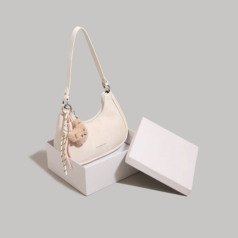 Mini crescent shoulder bag in cream with teddy bear charm and adjustable strap.