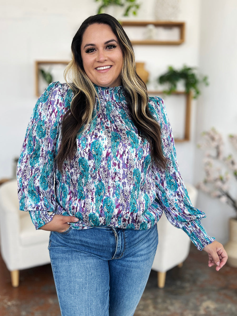 High neck abstract print blouse with smocked cuffs and hem in teal, purple, and white.