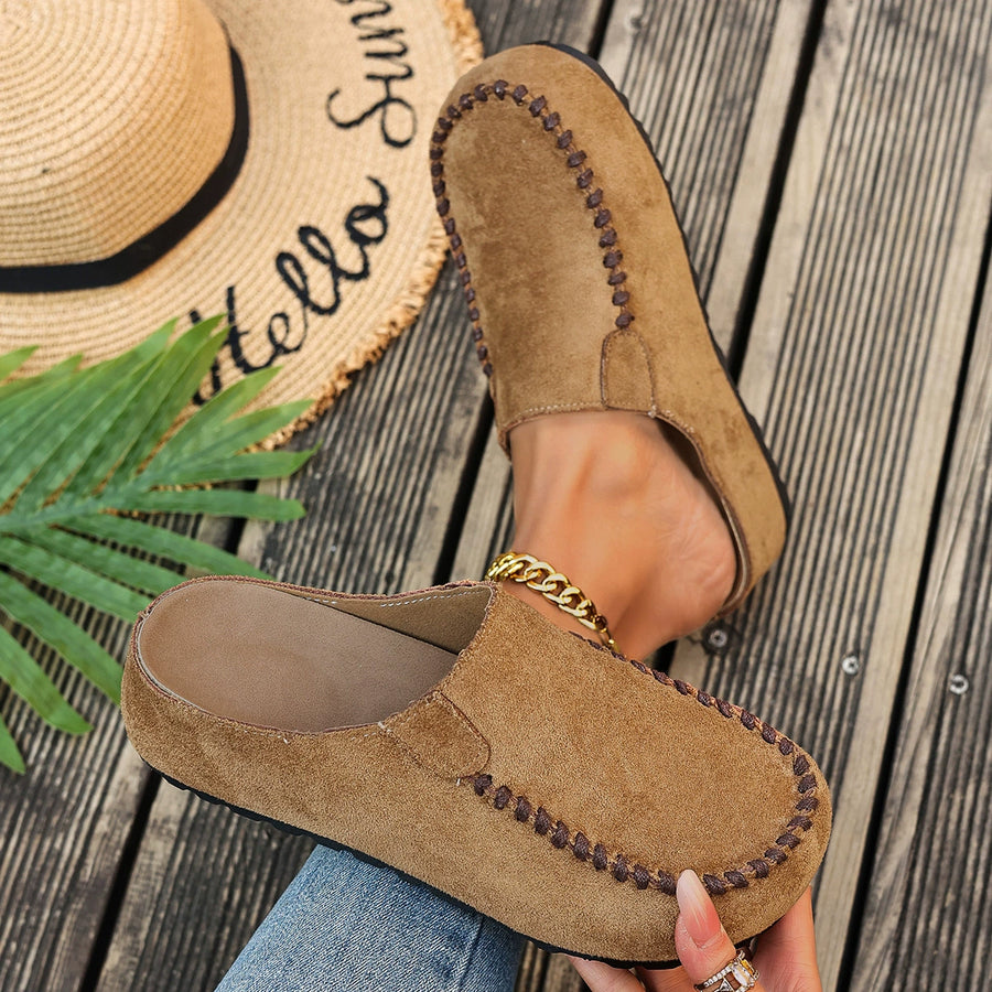 Brown suede slip-on flats with decorative stitching and a gold chain detail.