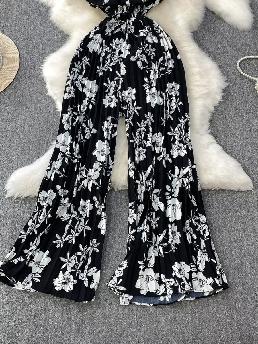 Black floral pleated wide-leg romper with a smocked waist and flowing legs.