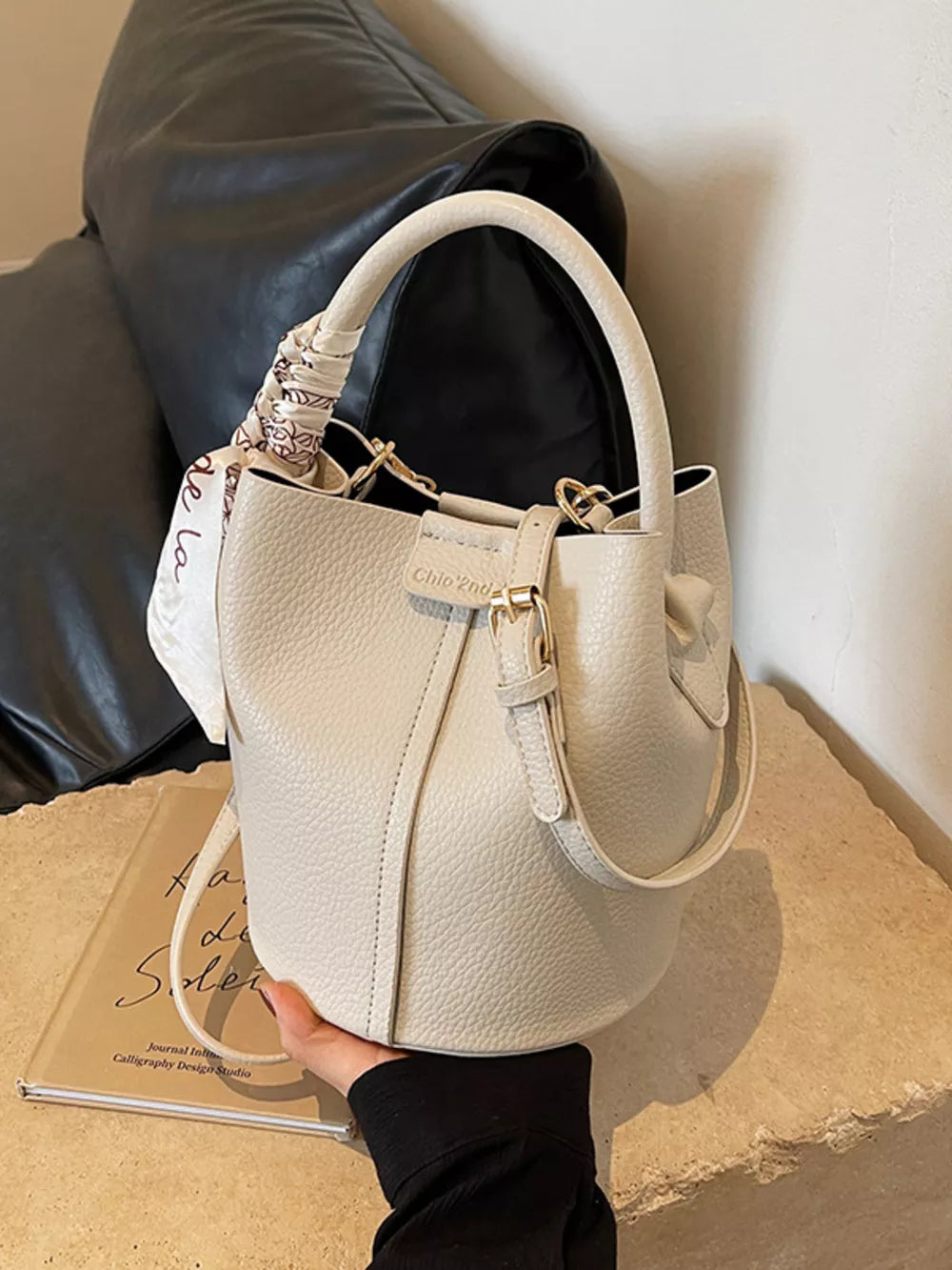 Beige pebbled leather bucket bag with top handle and decorative scarf detail.