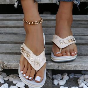 Beige perforated thong wedge sandals with gold embellishments and a chain ankle bracelet.
