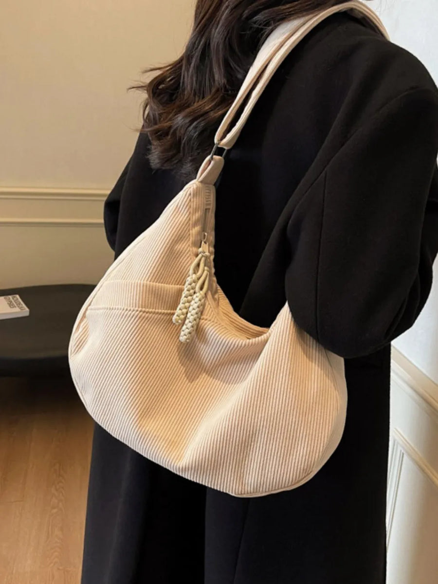 Beige corduroy crescent shoulder bag with tassel zipper pull and braided strap detail.