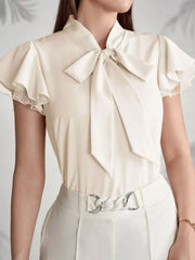 Beige tie-neck blouse with ruffle sleeves and lace trim, styled with white pants.