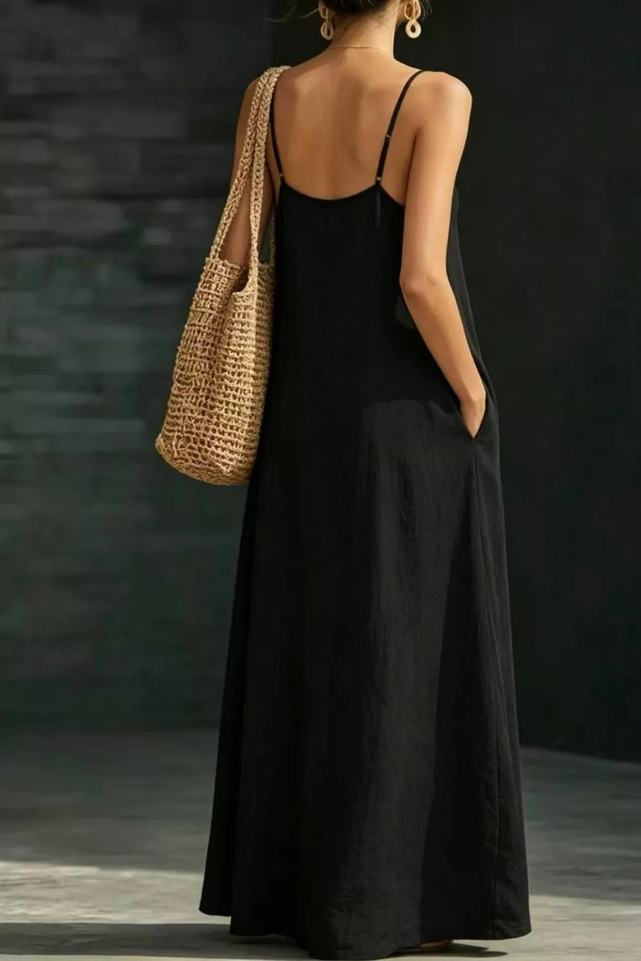 Black A-line maxi dress with spaghetti straps and pockets, worn with a straw bag.