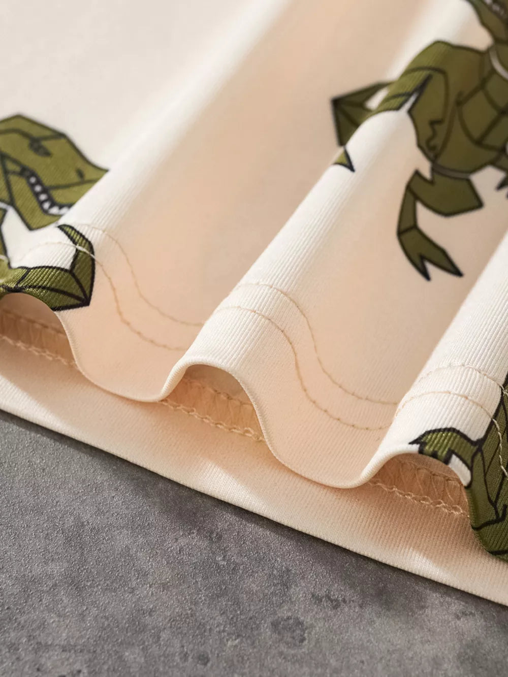 Cream cotton baby tee with green dinosaur print and stitched details.