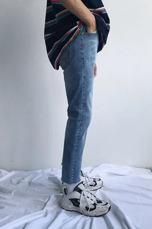 Medium wash denim jeans with raw hem and slim fit, styled with a striped shirt and sneakers.