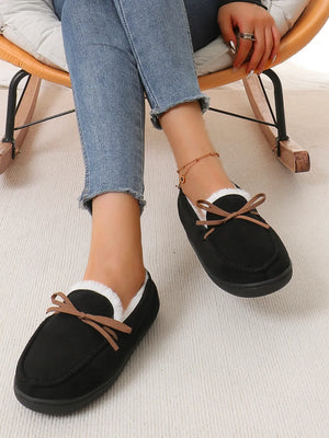 Black suede moccasin slippers with white faux fur lining and a brown bow detail.
