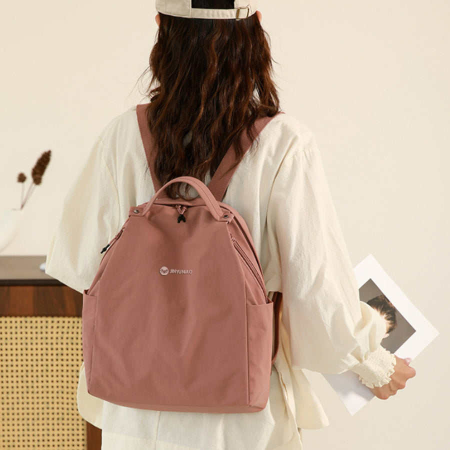 Blush pink nylon backpack with side pockets and top handles, worn by a woman.