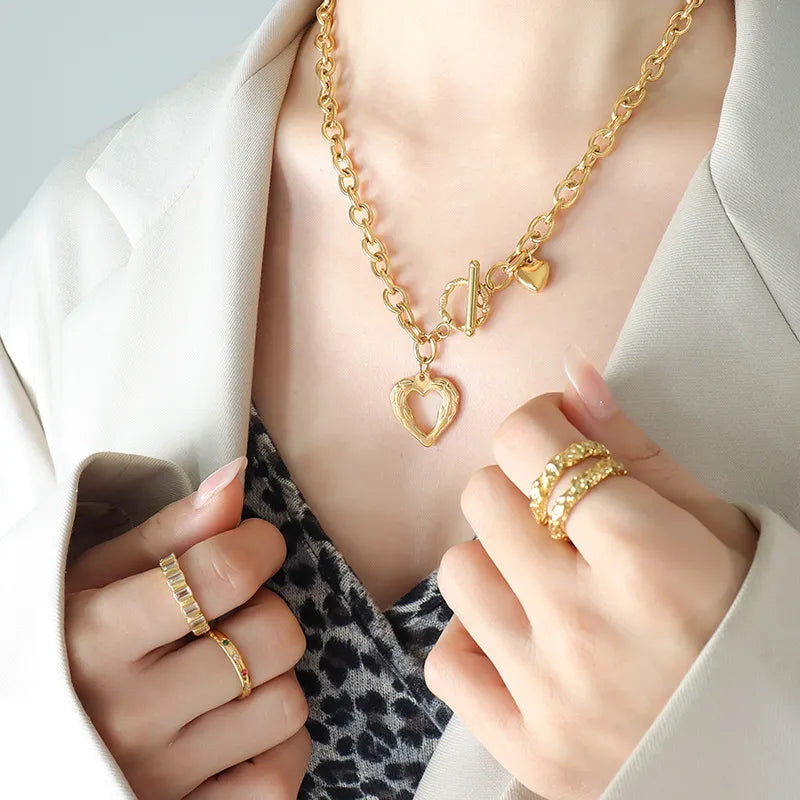 Gold open heart chain necklace with toggle clasp and charm, styled with rings.