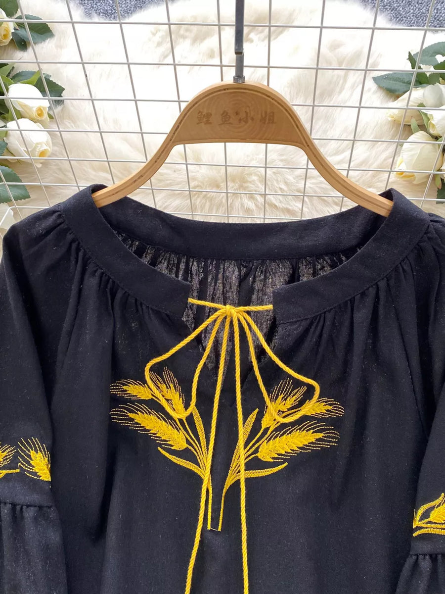 Black tie-neck blouse with yellow wheat embroidery and ruffled sleeves.