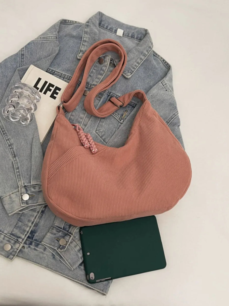 Pink corduroy crescent shoulder bag with braided tassel detail and adjustable strap.