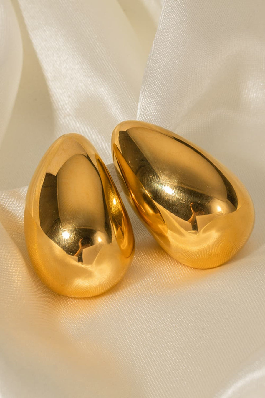 Gold teardrop stud earrings with a smooth, sculptural finish on satin fabric.