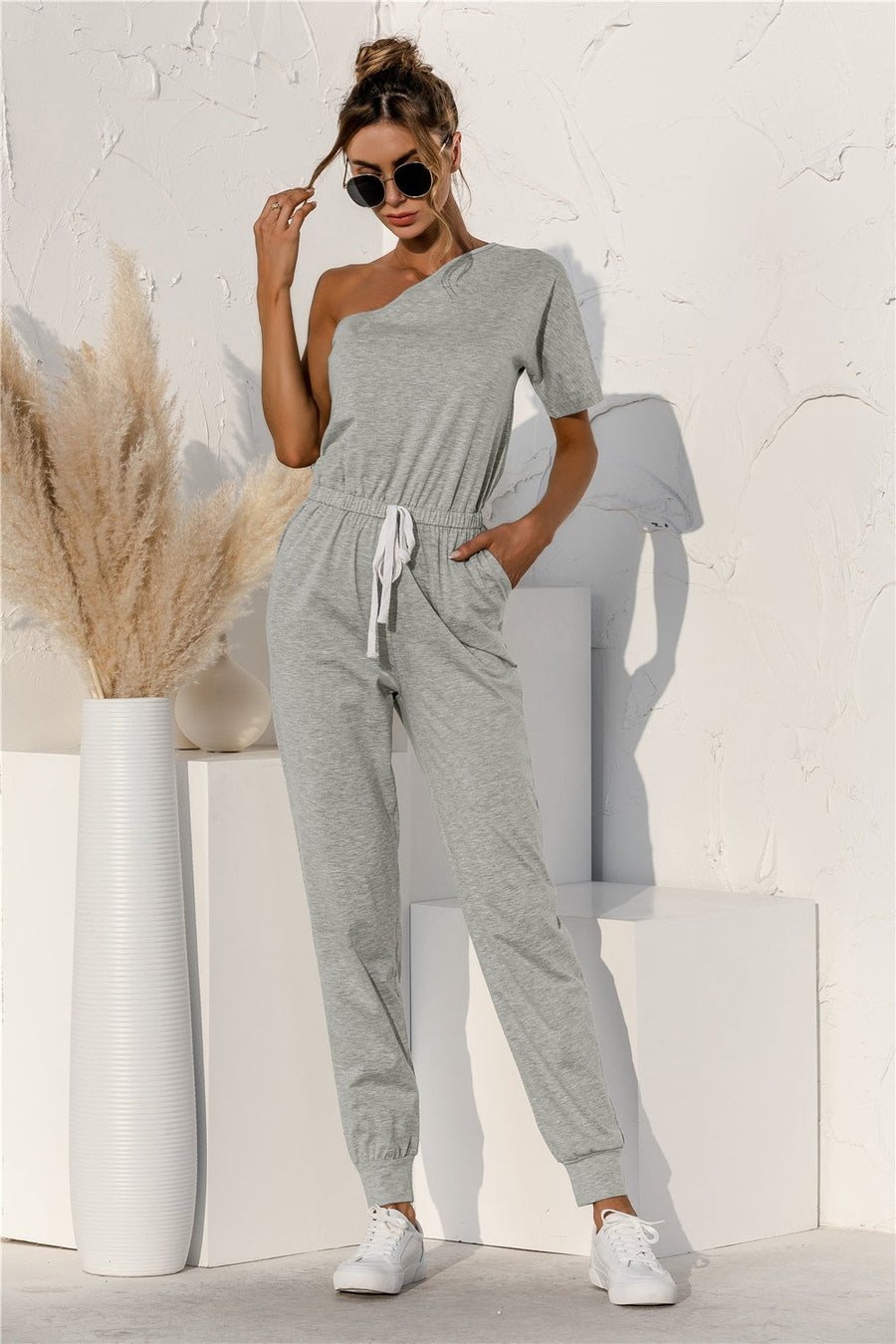 Gray asymmetric one-shoulder sweat jumpsuit with drawstring waist and cuffed jogger pants.