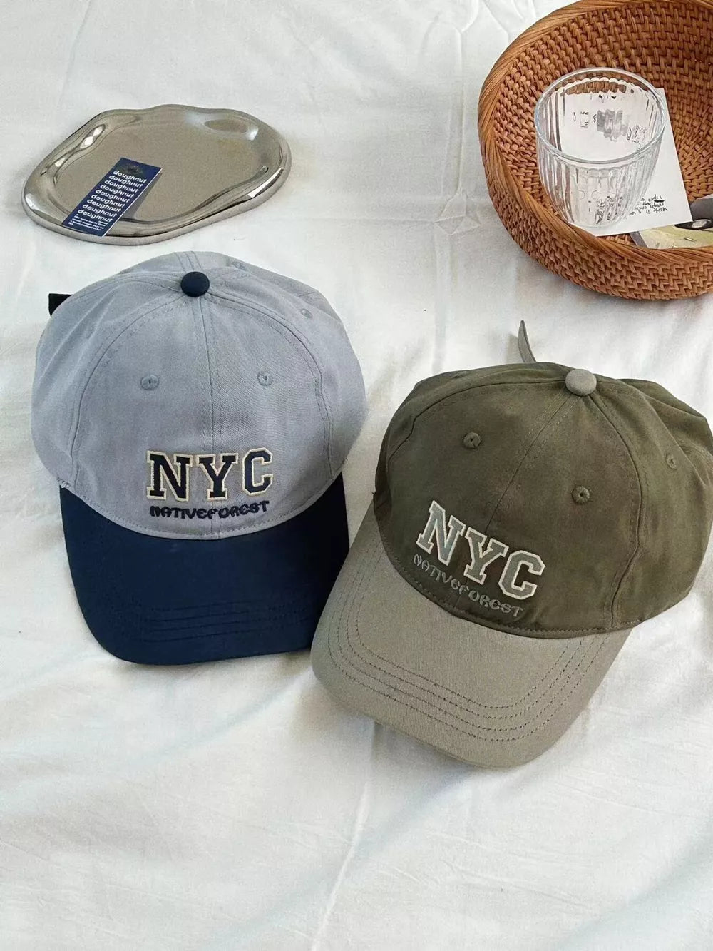 Cotton baseball caps with embroidered NYC detail, one grey with navy brim, one military green with grey brim.