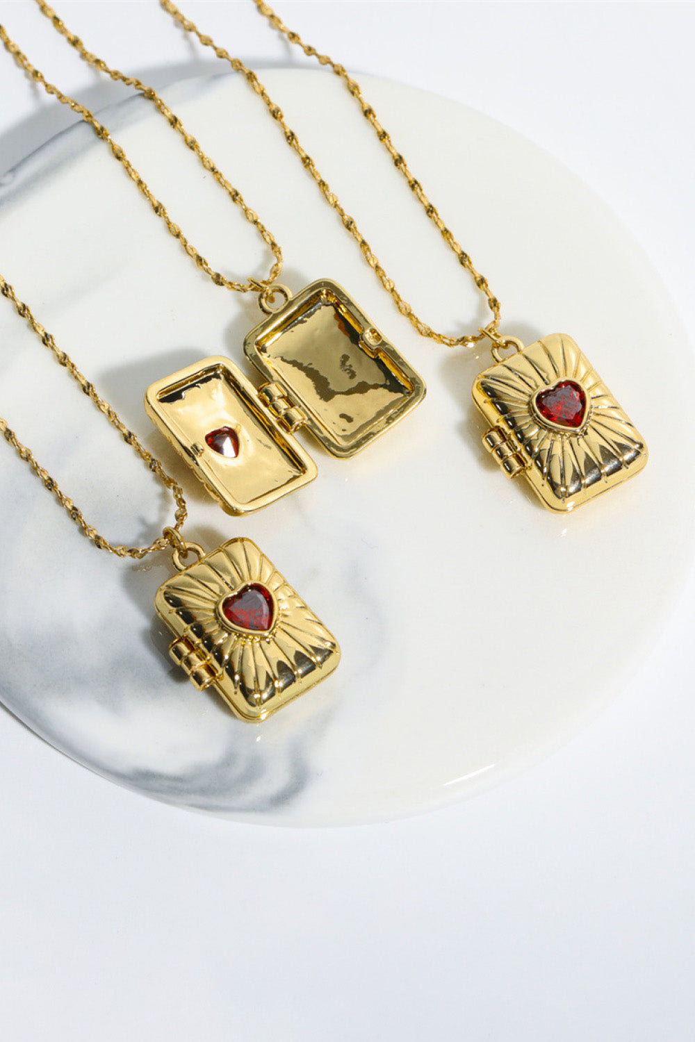 Gold locket pendant necklace with a red heart-shaped gem and sunburst detail.