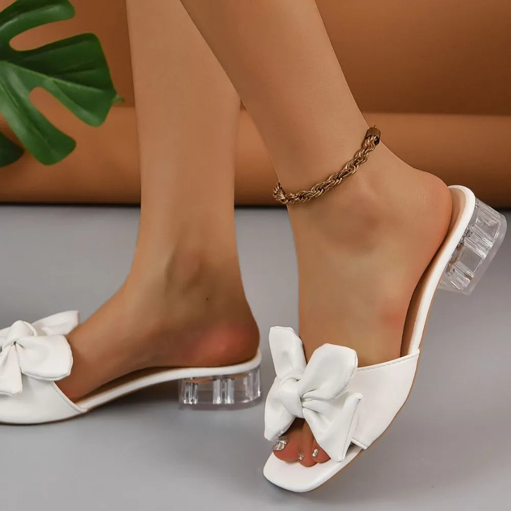 White bow slides with clear block heels and a gold chain anklet.