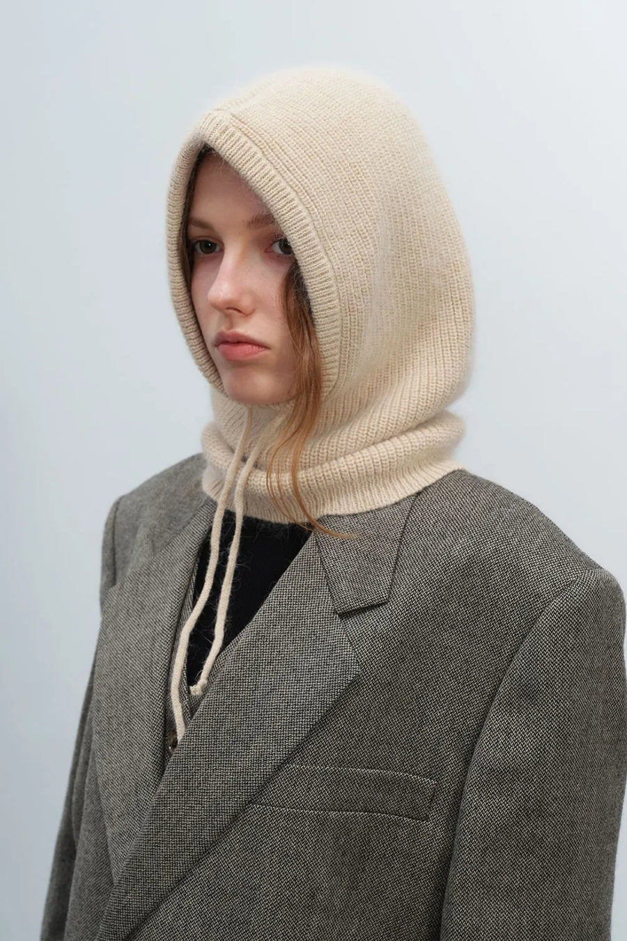 Beige knit balaclava with drawstring hood and ribbed trim, worn with a gray blazer.
