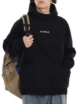 Black oversized hoodie with white embroidered text "Be Difficult." and a front pocket.