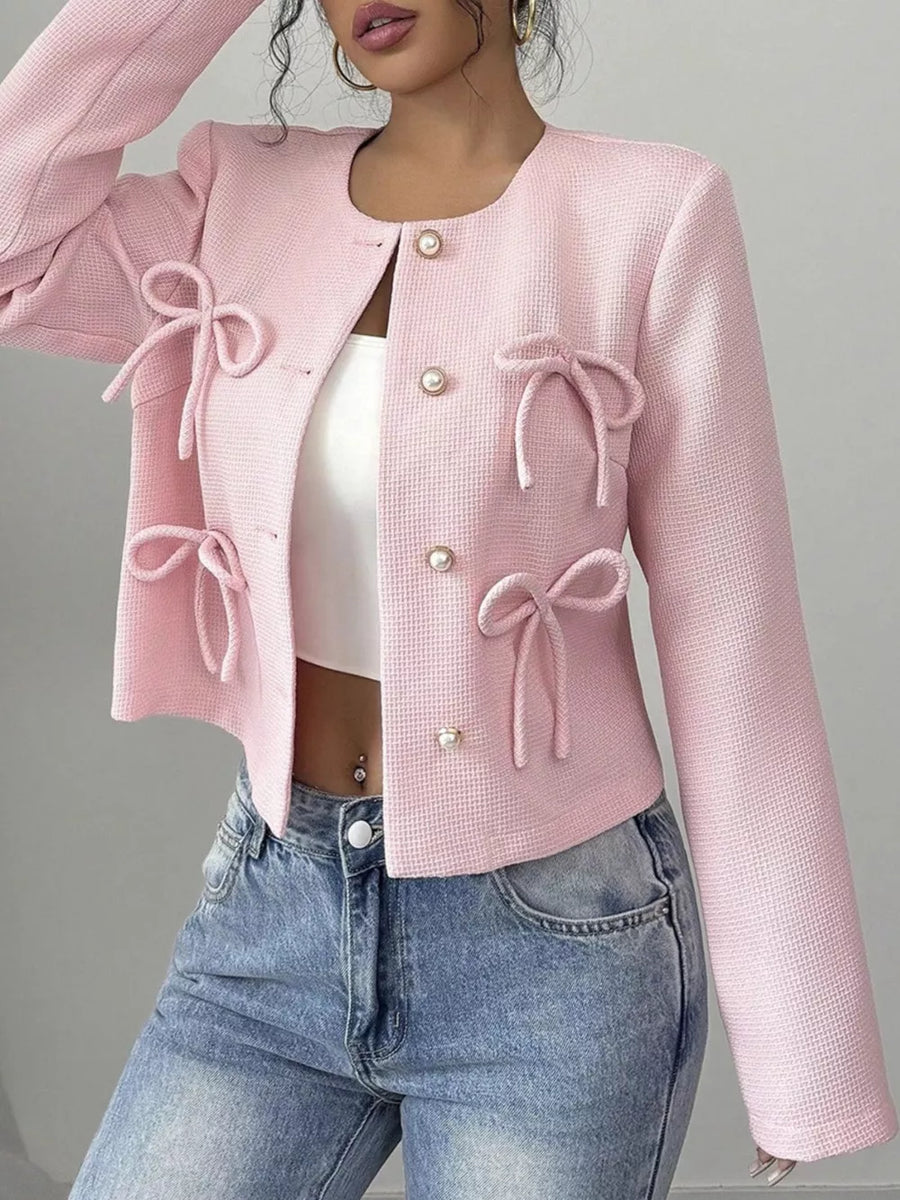 Pink textured jacket with pearl buttons and bow details, crew neck, long sleeves.