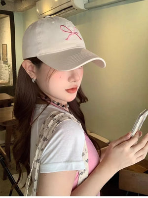 White baseball cap with pink bow embroidery, styled with a pink tank top and floral print shirt.