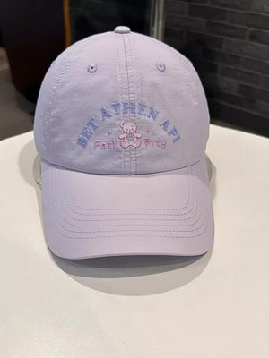 Lilac baseball cap with embroidered teddy bear and text detail.