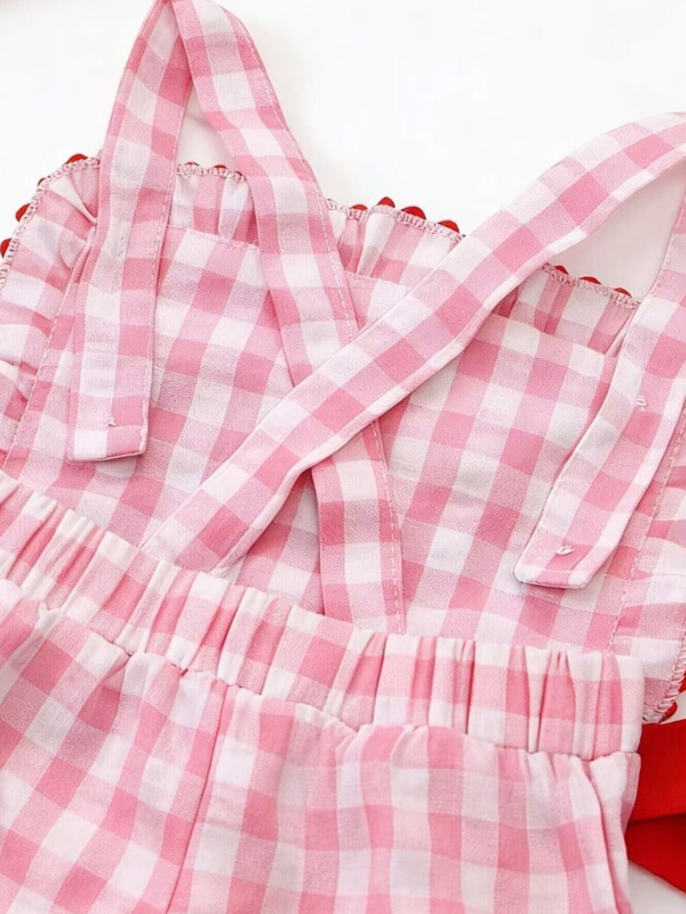 Blush pink gingham baby jumpsuit with criss-cross straps and red pom-pom trim.