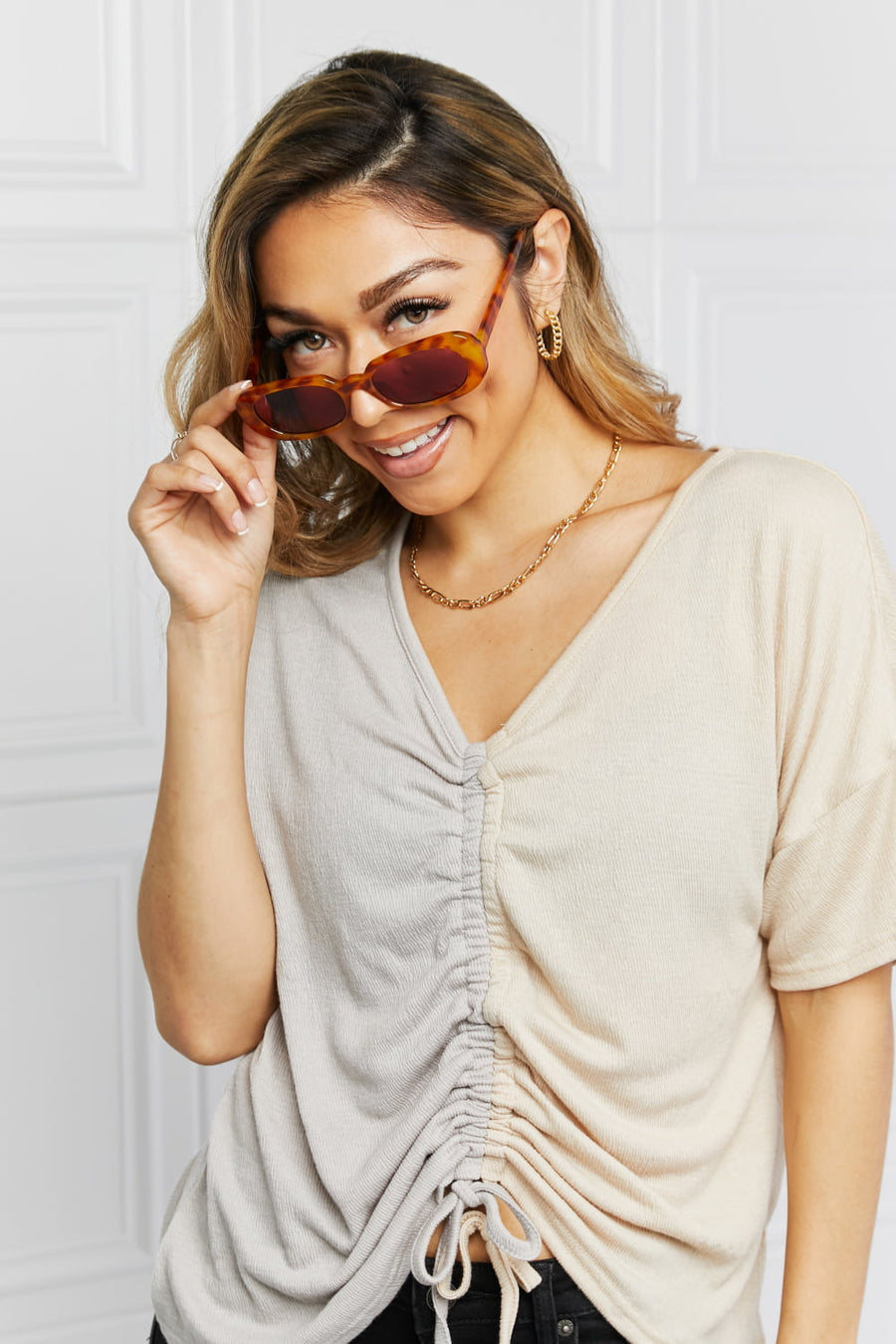 Tortoiseshell oval sunglasses with sunset lenses, held by a woman wearing a ruched two-tone top.