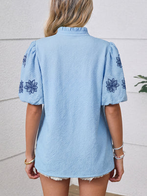 Light blue textured blouse with embroidered floral sleeves and ruffled mock neck.