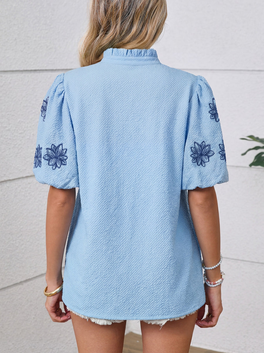 Light blue textured blouse with embroidered floral sleeves and ruffled mock neck.