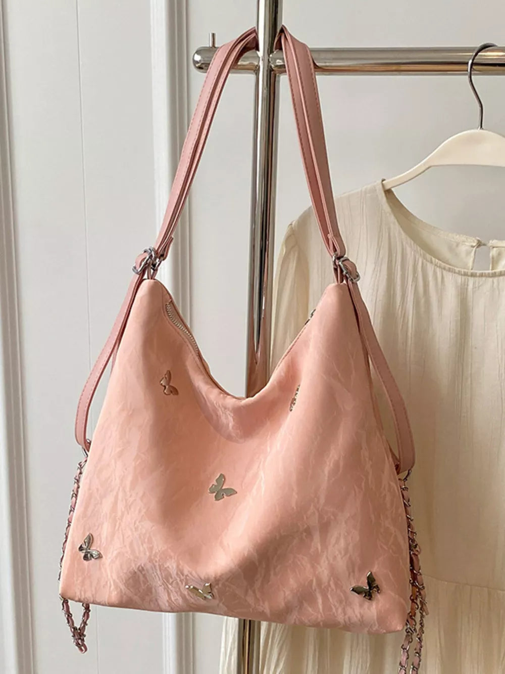 Blush pink shoulder bag with whimsical butterfly embellishments and chain strap detail.