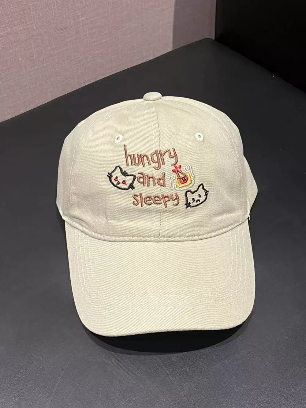 Beige baseball cap with "hungry and sleepy" text and embroidered cats.