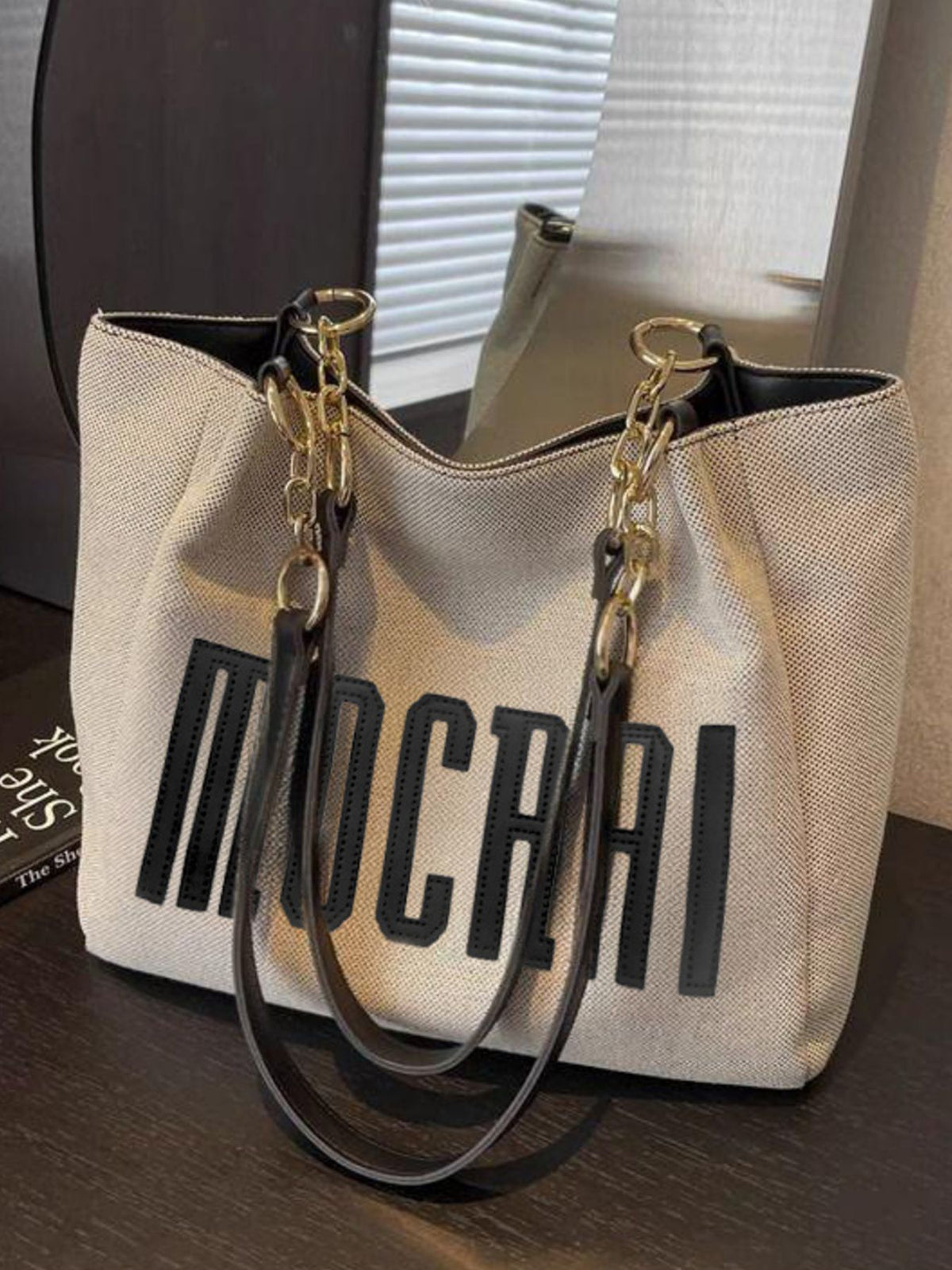 Textured canvas shoulder bag in beige with black lettering and gold chain accents.