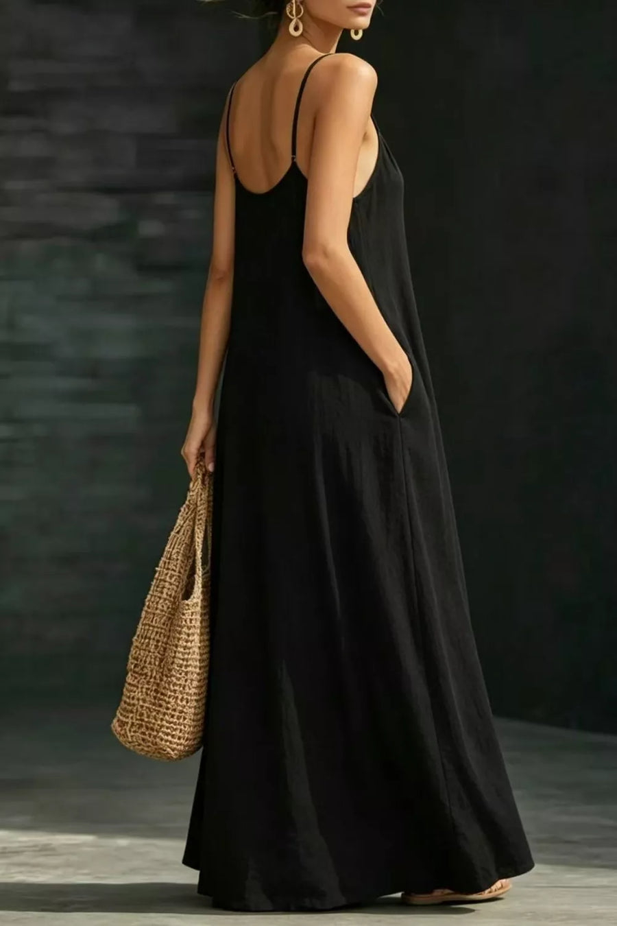 Black a-line maxi dress with spaghetti straps and pockets, held by a model with a straw bag.