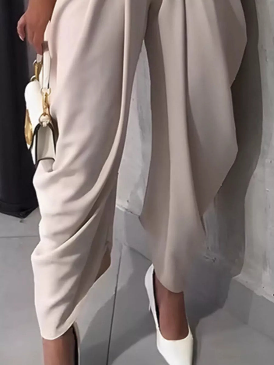 Light khaki sculptural pleated high-waisted pants with a buttoned closure and a white handbag.