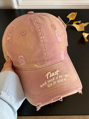 Distressed baseball cap in dirty pink with embroidered text and frayed details.