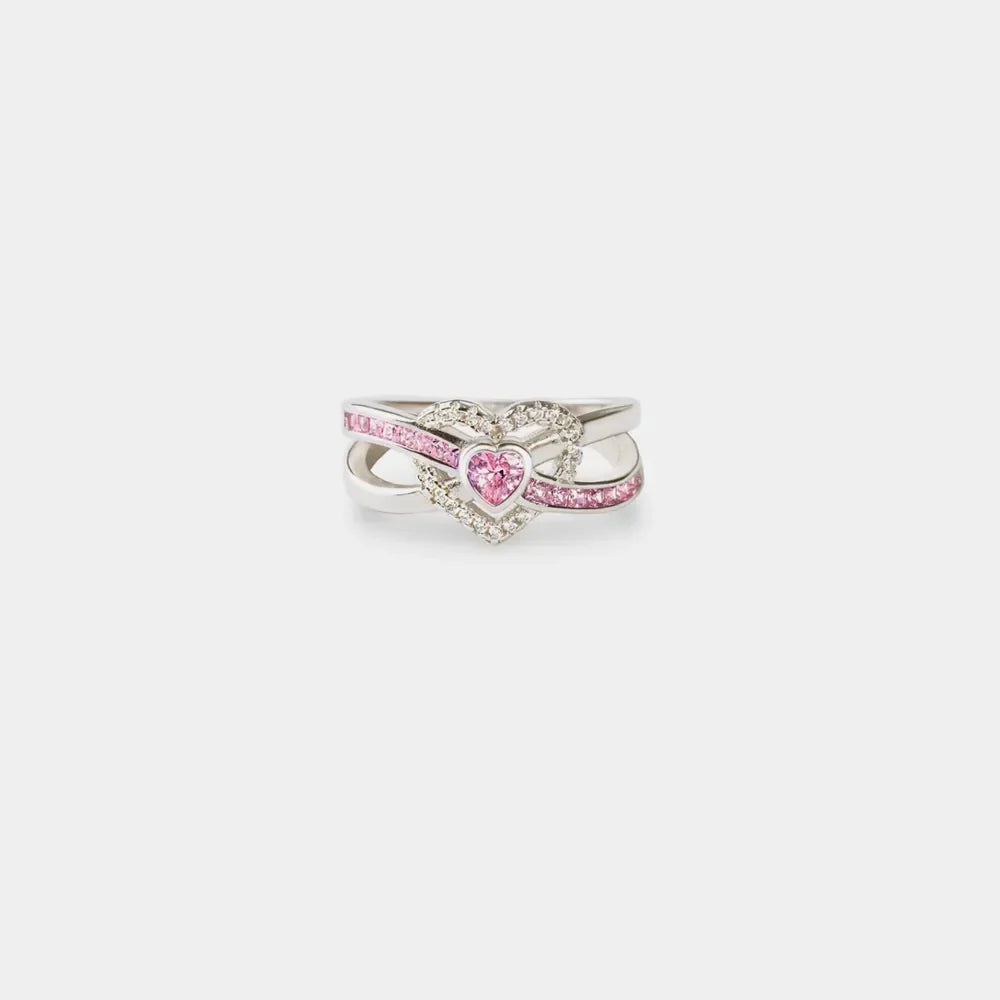 Silver ring with intertwined infinity band and a pink heart-shaped zirconia center surrounded by small clear stones.
